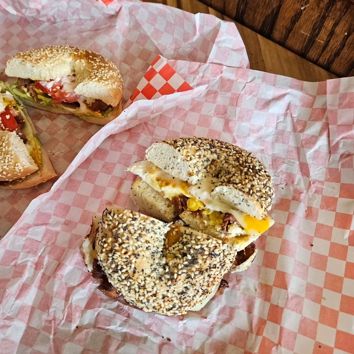 Two everything-bagel breakfast sandwiches with egg, cheddar and bacon on red gingham paper