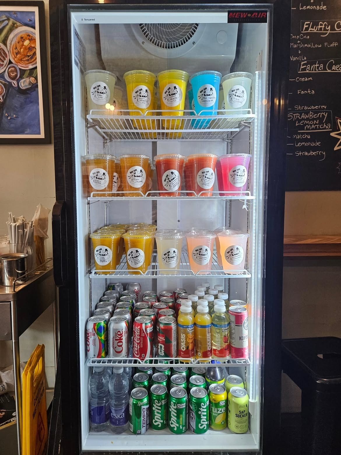 The drinks fridge: rows of homemade lemonades and iced teas in orange, yellow, blue and pink, each with a Rustic Cosmo sticker, cans below
